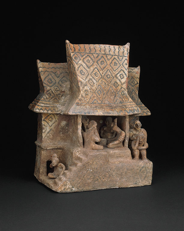 A small brown ceramic model of a house. The roof is steep & diamond-patterned, & the central room is open to reveal people sitting in a circle. Stairs descend from this floor to a small area with another sitting figure on the left, and two other figures are sitting to the right of the main room.